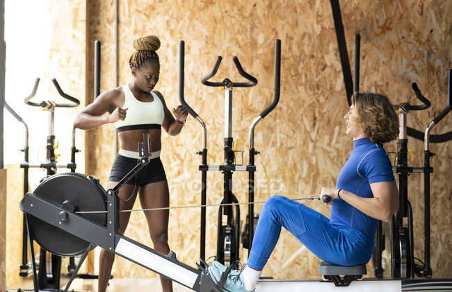 Atleta masculino sonriente haciendo ejercicio en el equipo de remo y hablando con una pareja étnica femenina mientras se miran en el gimnasio - foto de stock