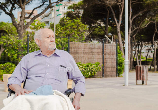 Senior male sitting in wheelchair in street on sunny day and looking away — Stock Photo