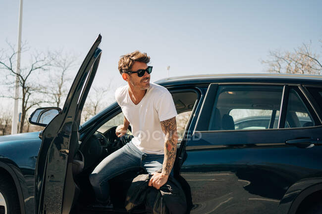 Веселий чоловічий водій в сонцезахисних окулярах виходить з сучасної машини, дивлячись в сонячний день — стокове фото