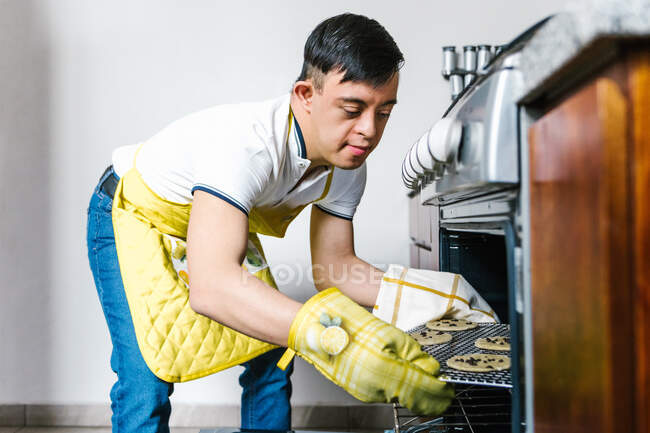 Ethnic teenage boy with Down syndrome putting raw chocolate chip cookies in oven while baking pastry in kitchen — Stock Photo