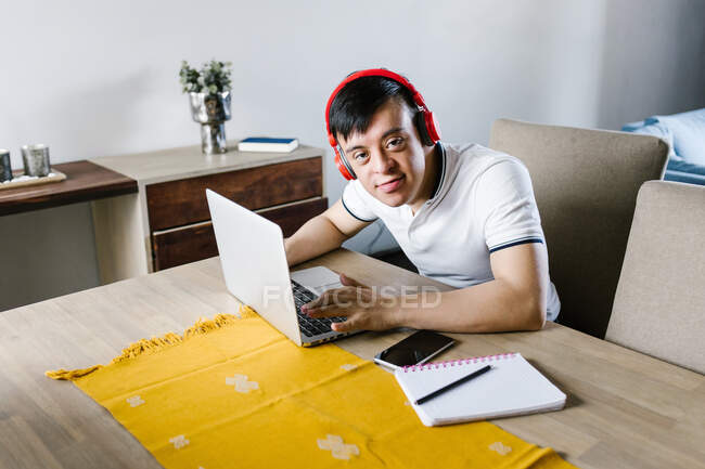 Alto ángulo de contenido latino adolescente chico con síndrome de Down netbook de navegación mientras está sentado en la mesa y estudiar en línea desde casa - foto de stock