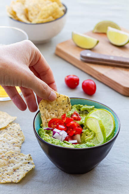 Crop unrecognizable person dipping cracker with herbs in delicious guacamole with fresh cherry tomato and lime slices — Stock Photo