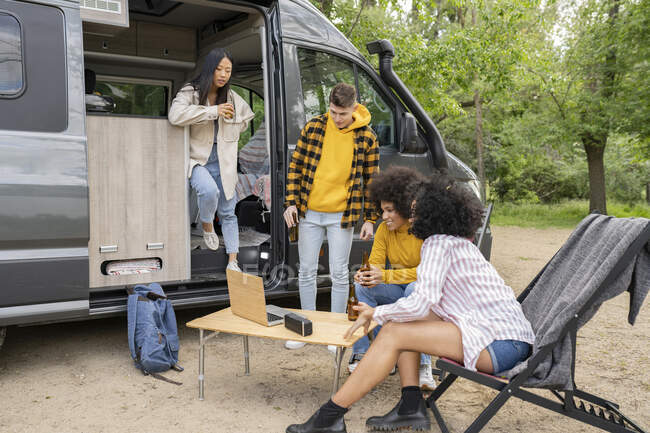 Group of multiracial travelers with beer gathering near van and watching video on netbook during road trip in countryside — Stock Photo