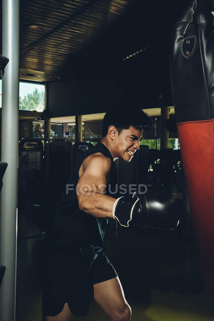 Giovane focalizzato asiatico uomo formazione pugilato esecuzione pugni mentre si esercita con pesante sacco da boxe in una palestra moderna — Foto stock