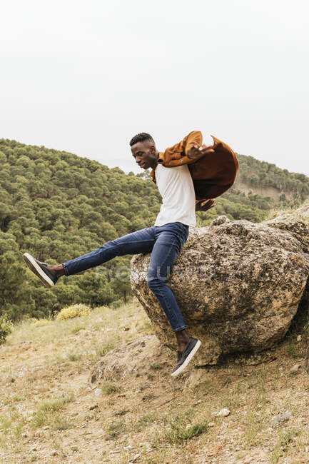 Trendy African American male model in fashionable outfit jumping from stone in highlands — Stock Photo
