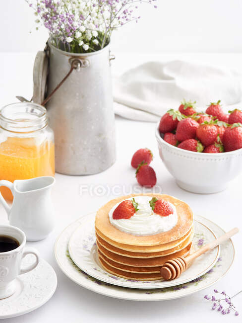 Elegantes platos de mesa con incluso una pila de deliciosos panqueques rematados con crema y fresas de verano para el desayuno - foto de stock