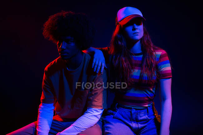 Corpo pieno di auto assicurato giovane donna in abito alla moda seduto sulla sedia con mano sulla spalla del fidanzato afroamericano in studio scuro in luci al neon — Foto stock