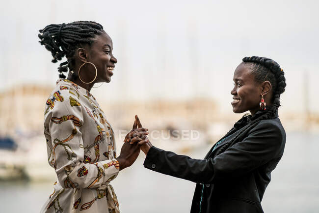Модные хитроумные улыбчивые афроамериканские дамы проводят время вместе и сжимают руки в парке в яркий день — стоковое фото