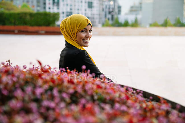 Vista lateral de la alegre mujer musulmana en pañuelo para la cabeza sentada en el banco cerca del parterre en la calle y mirando a la cámara - foto de stock