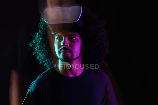 Delizioso maschio latino con capelli ricci e moderni occhiali di realtà virtuale in piedi in studio su sfondo nero con luci al neon — Foto stock