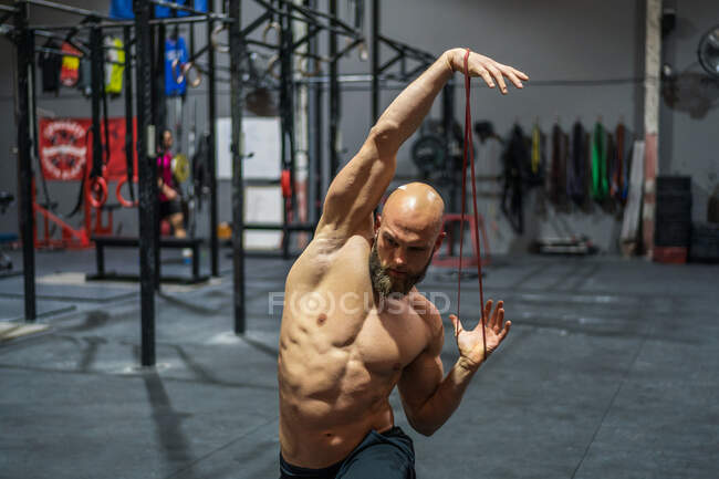 Hemdloser bärtiger Mann schaut beim funktionellen Training im Fitnessstudio weg und streckt das Gummiband mit den Armen — Stockfoto