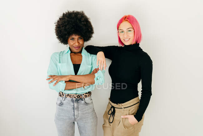 Веселые молодые розовые волосы женщина и афроамериканская девушка в стильном наряде, веселясь вместе на белом фоне — стоковое фото