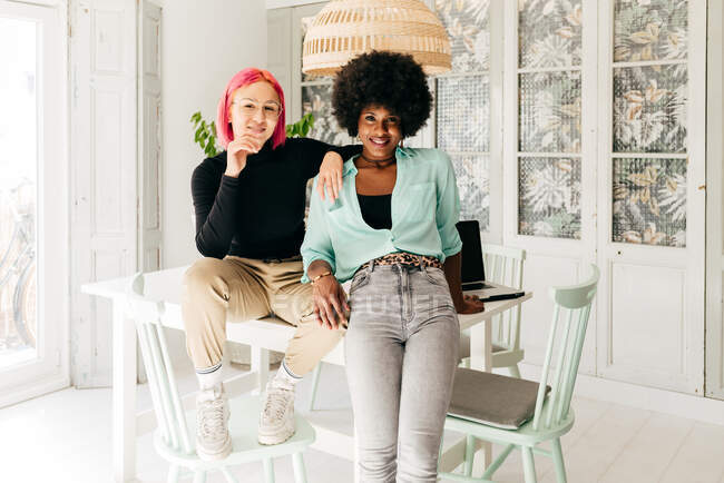 Fröhliche junge stilvolle multirassische Freundinnen blicken in die Kamera, während sie zusammen auf einem Tisch im hellen Raum sitzen — Stockfoto