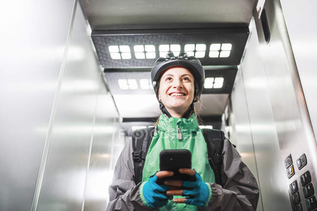 Bajo ángulo de mensajería femenina alegre en uniforme y casco sonriendo y navegando por el teléfono celular en el ascensor mientras hace la entrega - foto de stock