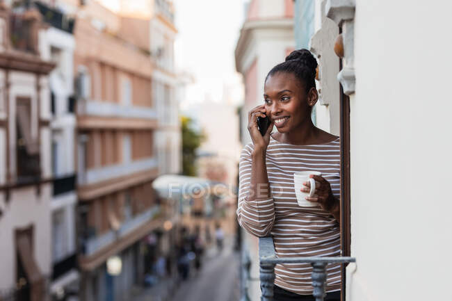 Afroamerikanerin mit Tasse Heißgetränk telefoniert auf Balkon — Stockfoto