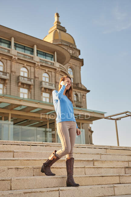 Полное тело улыбающейся взрослой леди в повседневной одежде с телефонным звонком во время прогулки по лестнице в районе города рядом со старым зданием при дневном свете — стоковое фото