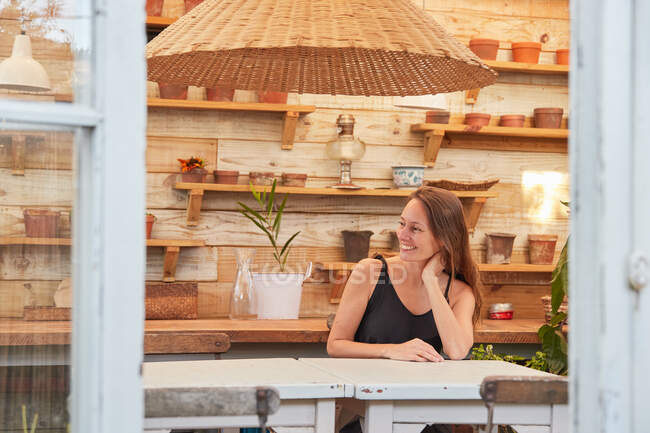 Jardinero femenino positivo sentado en la mesa de madera en el invernadero con plantas en maceta y mirando hacia otro lado - foto de stock