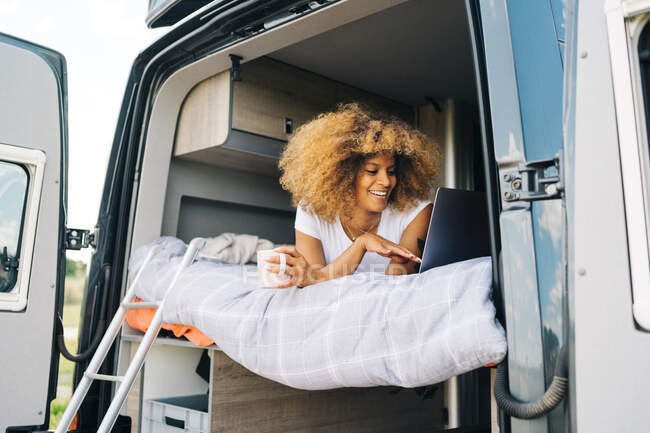 Optimistic black woman with mug of hot drink smiling and browsing social media on netbook while lying on bed in RV and resting in morning — Stock Photo