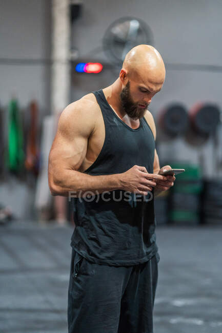 Вид сбоку мышечного лысого спортсмена, стоящего в современном тренажерном зале, просматривающего смартфон во время перерыва на тренировку — стоковое фото