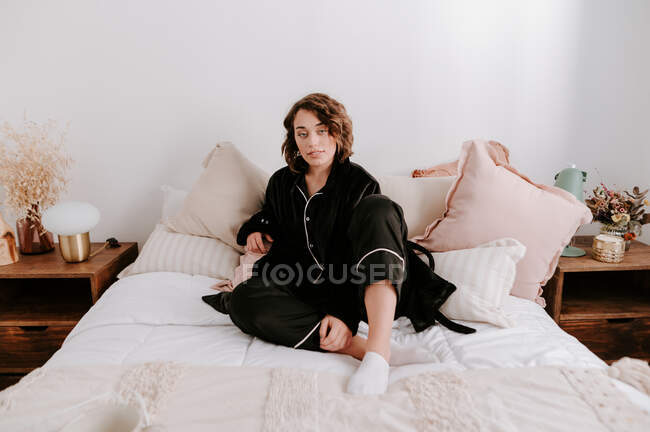 Ніжна жінка в чорній піжамі розслабляється на м'якому ліжку в спальні вранці і дивиться на камеру — стокове фото