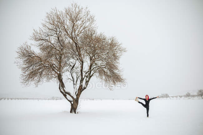 Удаленный взгляд на гибкую женщину, практикующую йогу в Уттхите Hasta Padangushthasana, стоя на снежном поле зимой — стоковое фото