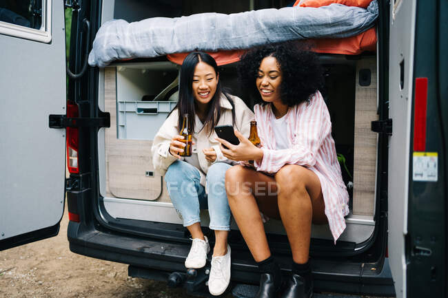 Mujeres jóvenes multiraciales con cerveza navegando en el teléfono inteligente mientras están sentadas en la camioneta durante el viaje por carretera - foto de stock