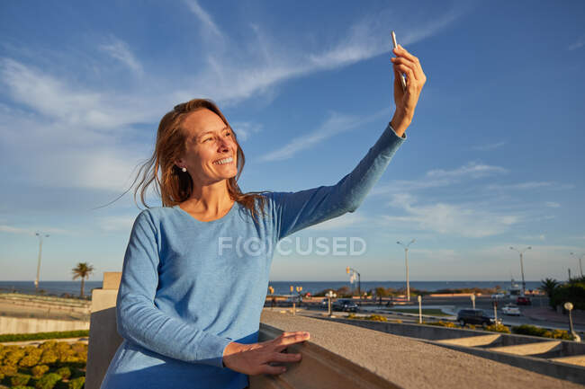 Улыбающаяся взрослая женщина делает селфи, опираясь на забор возле океана на городской улице в солнечный день — стоковое фото