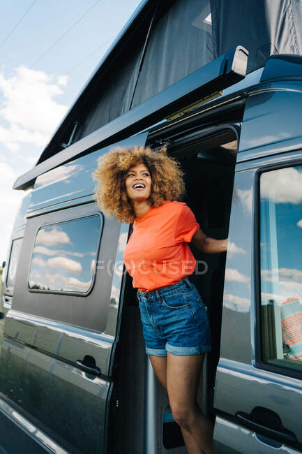 Happy African American woman with curly hair admiring countryside while standing behind opened door of RV during road trip in summer — Stock Photo