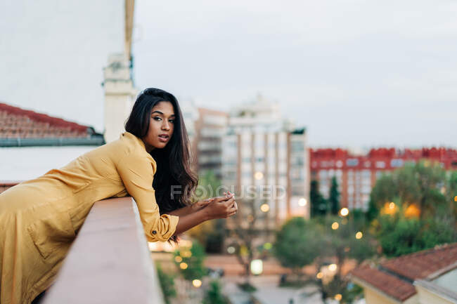 Junge hispanische Frau in lässiger Kleidung lehnt am Geländer und blickt in die Kamera, während sie es sich abends auf dem Balkon in der Stadt gemütlich macht — Stockfoto