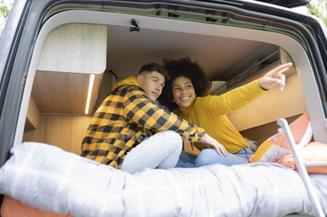 Low angle of African American woman smiling and pointing away for young boyfriend while sitting cross legged on bed in van during road trip — Stock Photo
