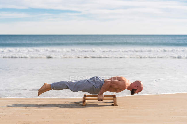 Боковой вид лысого сильного спортсмена, делающего параллельную стойку на руках с барами, работающими на песчаном побережье — стоковое фото