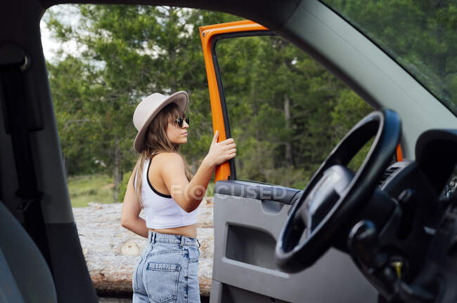 Vista lateral del explorador femenino de pie cerca de la puerta de la camioneta abierta estacionada en la naturaleza en verano - foto de stock