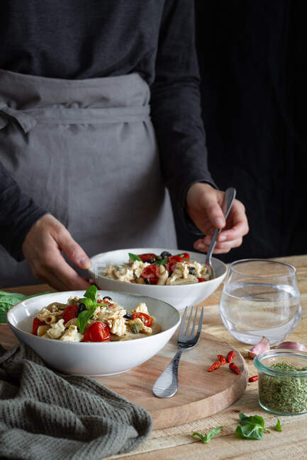 Cultivo pessoa anônima cozinhar saborosa massa assada com metades de tomate cereja e folhas de manjericão fresco em tigelas — Fotografia de Stock