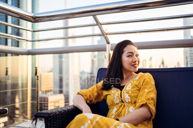 Young female traveler sitting on rooftop terrace of modern coffee shop at sunset in Dubai city and looking away — Stock Photo