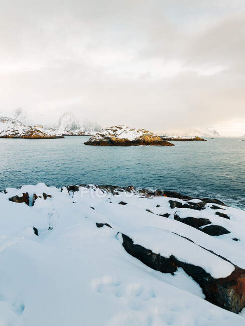Falésias nevadas viradas para ilhotas no meio de um mar ondulado com céu nublado no dia de inverno nas Ilhas Lofoten, Noruega — Fotografia de Stock