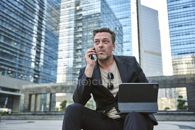 Homme d'affaires ayant un appel sur son smartphone et travaillant avec sa tablette tout en étant assis autour des bâtiments du bureau — Photo de stock