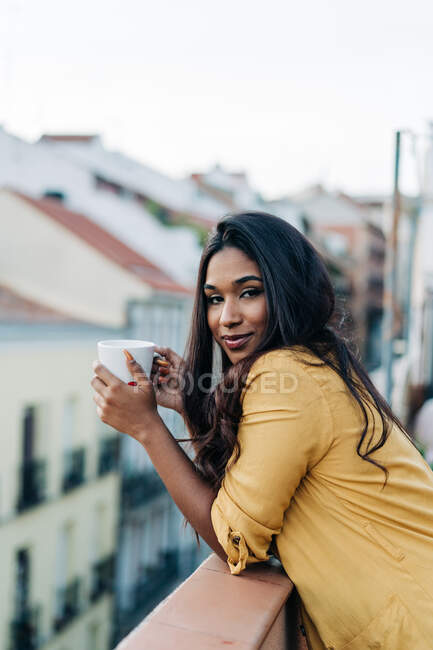 Seitenansicht einer jungen hispanischen Frau, die in die Kamera blickt und Heißgetränk genießt, während sie sich abends auf dem Balkon ausruht — Stockfoto