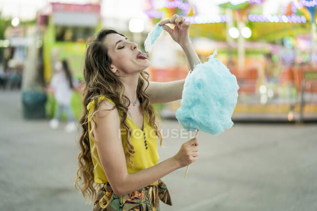 Вид сбоку на детскую женщину, которая ест сладкий голубой хлопок, веселясь и наслаждаясь выходными на ярмарке летом — стоковое фото