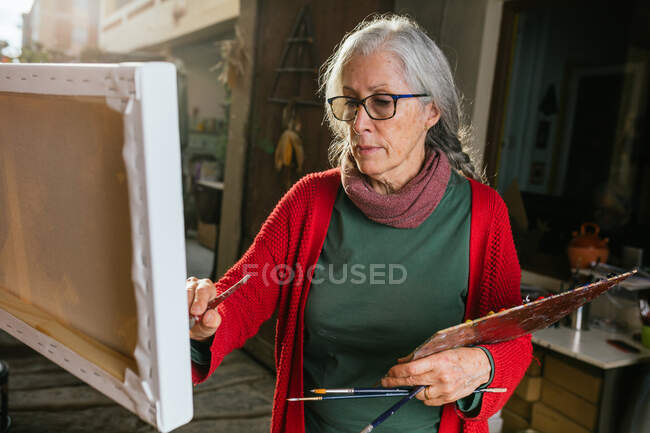 Aged female artists in protective mask painting on canvas in backyard on sunny day in summer — Stock Photo