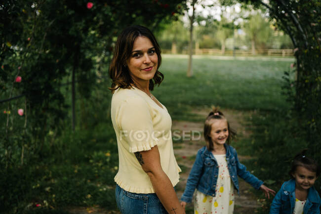 Молода жінка з чарівними маленькими доньками в подібних вбраннях, ходячи разом на зеленому трав'янистому газоні в літньому парку — стокове фото