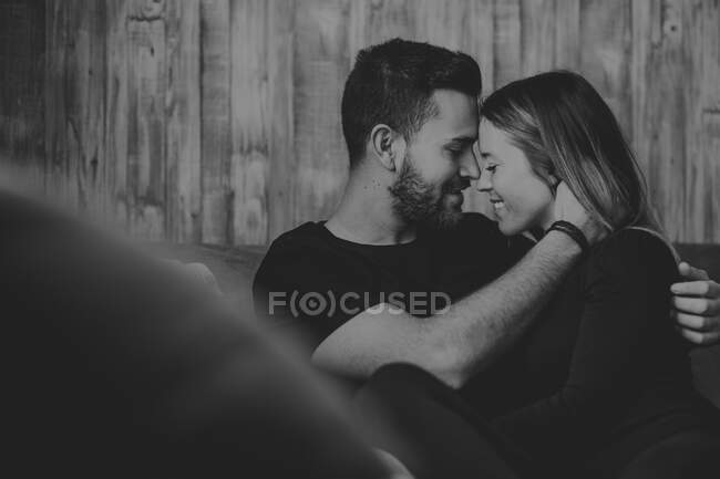 Mujer sonriente abrazando y besando a un hombre alegre en la frente mientras están sentados en un cómodo sofá en casa juntos - foto de stock