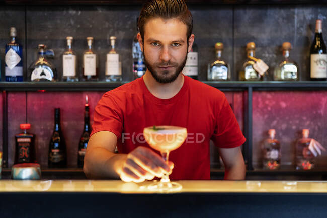 Cheerful barman serving glass with alcohol cocktail on counter in bar and looking at camera — Stock Photo