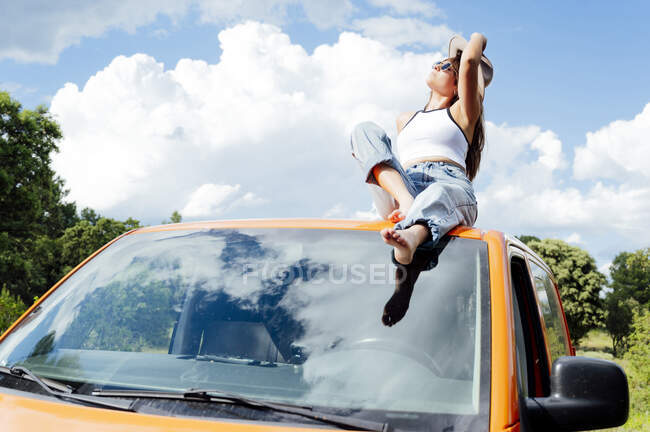 Спокойная женщина-исследователь сидит на крыше фургона и наслаждается летним приключением в солнечный день — стоковое фото