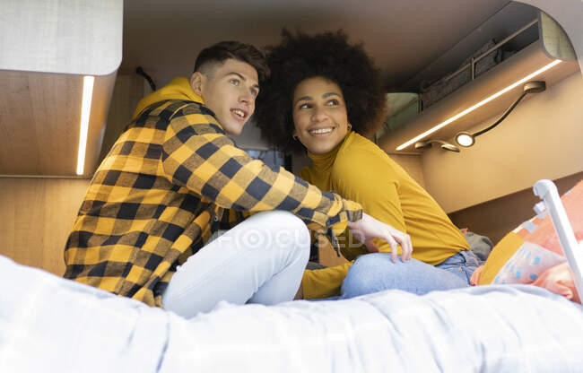 Low angle of African American woman smiling with young boyfriend while sitting cross legged on bed in van during road trip looking away — Stock Photo