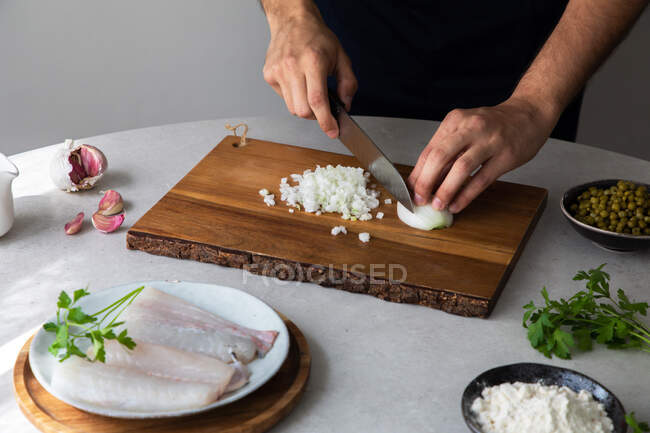 Hombre irreconocible cocinero cortando cebollas con cuchillo en una tabla de madera cerca de guisantes y filete de merluza con hierbas durante la preparación de alimentos en la cocina - foto de stock