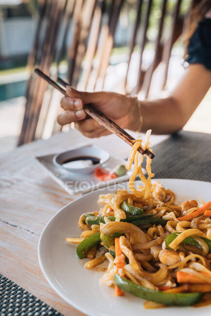 Anonymous female tourist with delicious pasta between food sticks above table with soy sauce and pickled ginger slices outdoors — Stock Photo