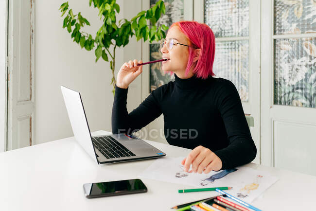 Задумчивая женщина-фрилансер в повседневной одежде и очках работает за столом с ноутбуком и рисует модные эскизы с карандашами — стоковое фото