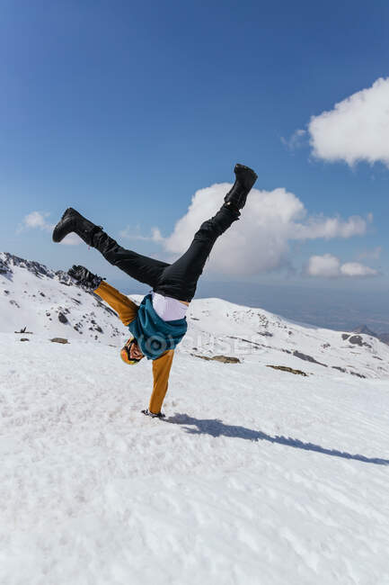 Anonymous sportsman in goggles standing on hand with raised legs on snow against Sierra Nevada under cloudy blue sky — Stock Photo