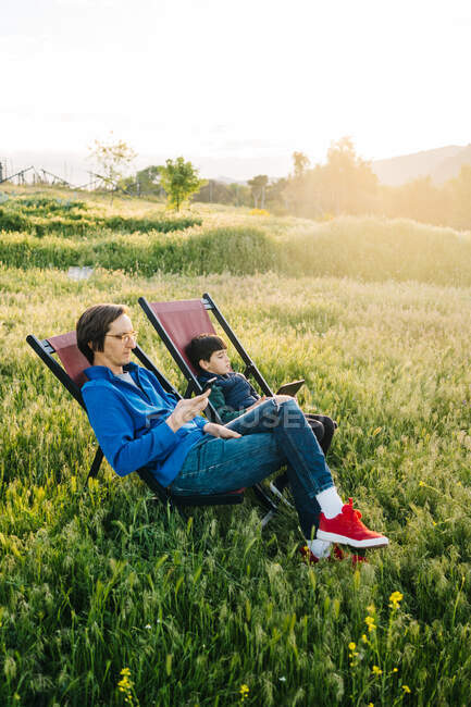Vista lateral do pai e do filho relaxando em redes dobráveis no meio da montanha ao pôr do sol — Fotografia de Stock