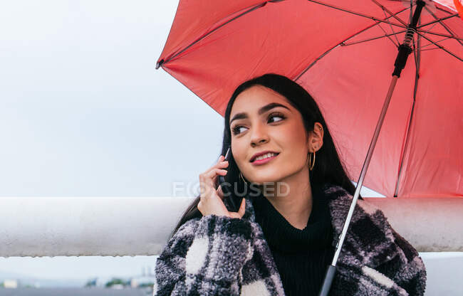 Этническая женщина в клетчатом пальто просматривает мобильный телефон, стоя под красным зонтиком в дождливый день на мокрой улице — стоковое фото